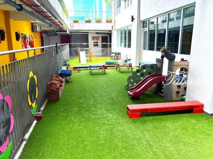 Bright outdoor playground with artificial grass, colorful play equipment, and wooden climbing structures in a modern building courtyard.