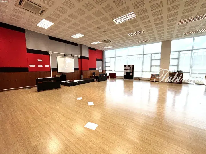 Spacious hall with wooden flooring, red and grey walls, projector screen, and large windows letting in natural light.