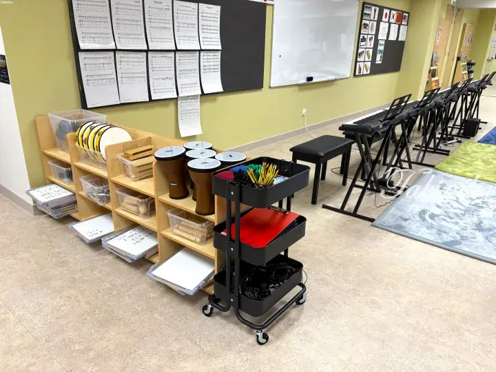 Music classroom with keyboards, percussion instruments, and sheet music displayed on the wall.