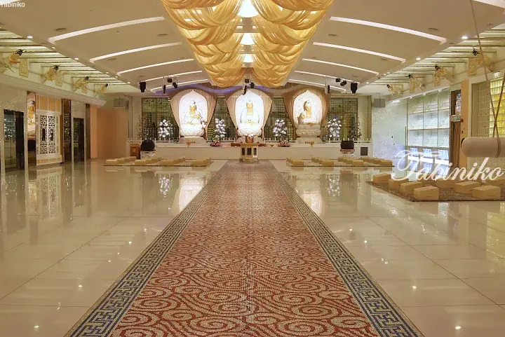 Spacious hall with three illuminated Buddha statues, golden ceiling drapes, and a patterned carpet runner.
