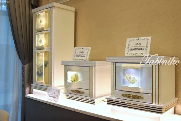 Display of elegant cremation urn compartments featuring glass doors, floral motifs, and gold accents with model labels.