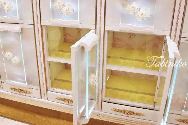 Luxurious white display cabinet with gold accents and floral glass doors, featuring yellow shelves inside.