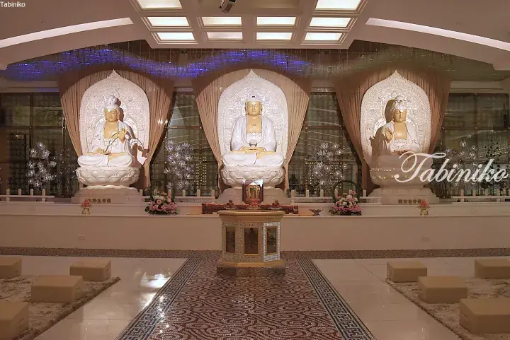 Three white Buddha statues in a serene prayer hall with ornate halos, floral offerings, and meditation cushions.