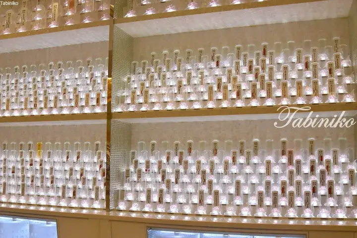 Rows of illuminated glass memorial tubes with pink lotus bases and Japanese text displayed in a modern Buddhist temple.