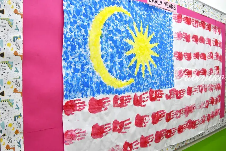 Children&rsquo;s handprint art depicting the Malaysian flag with blue, yellow crescent and star, and red stripes on a school bulletin board.