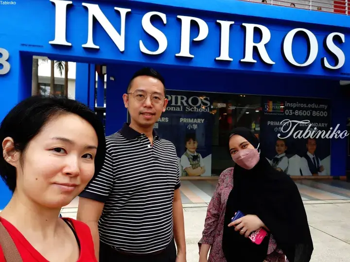 Three people posing for a selfie in front of the blue Inspiros International School building in Malaysia.