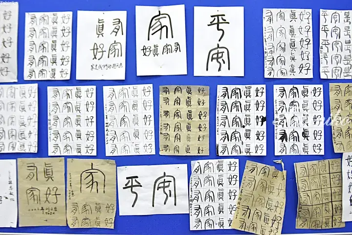 Blue bulletin board displaying handwritten Chinese calligraphy papers featuring characters for &lsquo;home&rsquo; and &lsquo;peace&rsquo;.