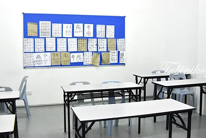 Empty classroom with white tables, blue chairs, and a bulletin board displaying student calligraphy practice sheets.