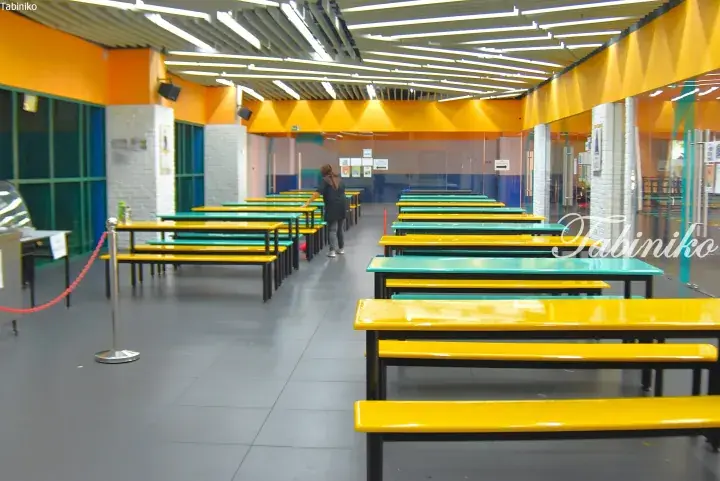 Bright school cafeteria with yellow and teal tables and benches, featuring modern ceiling lights and orange walls.