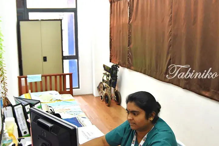 A caregiver in green scrubs works at a computer in a patient room with a bed and wheelchair.