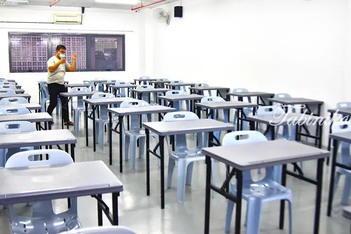Man in mask gesturing in empty classroom with rows of tables and blue chairs, likely a training center or school.