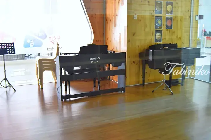A Casio Privia digital piano and a grand piano in a bright music room with wooden floors and walls.