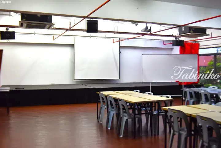 Empty event hall with wooden flooring, projector screen, whiteboard, and tables set up for a presentation or meeting.
