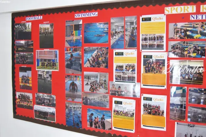 Red school bulletin board displaying photos of football, swimming, and netball teams alongside The Quadlet magazine articles.