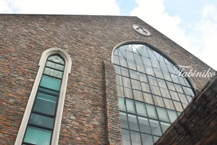 Close-up of a building facade featuring textured brown stone walls, a tall arched window, and a large grid-patterned glass window.