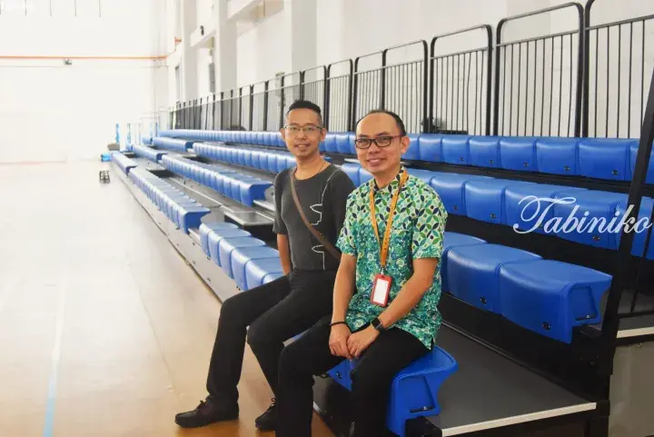 Two men sitting on blue retractable bleachers inside a modern indoor sports hall.