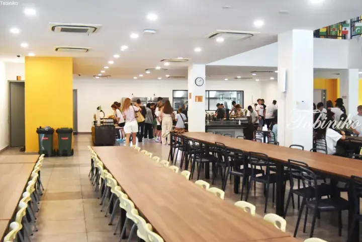 Bright school cafeteria with long wooden tables, students lining up at the food counter, and yellow accent walls.