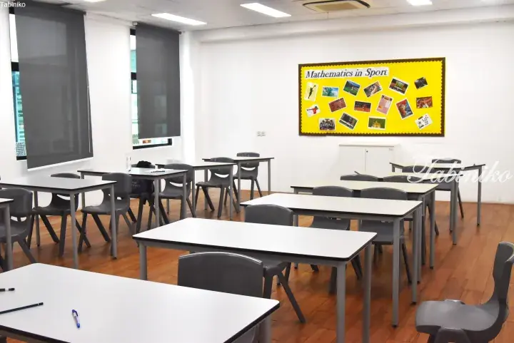 Bright classroom with white desks, grey chairs, and a yellow bulletin board displaying &lsquo;Mathematics in Sport&rsquo; with sports photos.