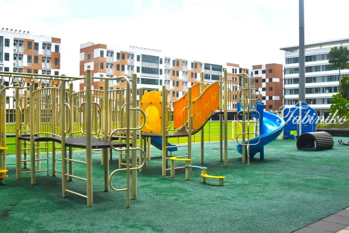 Colorful playground equipment with slides and climbing structures on green safety flooring in front of modern residential buildings.