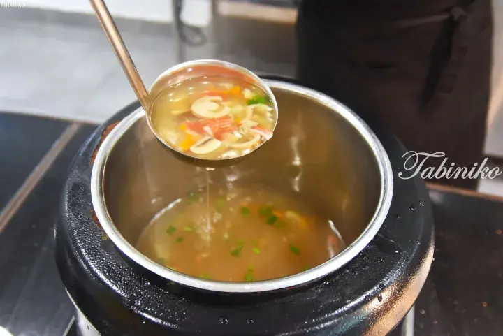 A ladle scoops clear soup with mushrooms and vegetables from a large black pot.