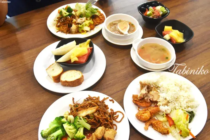 A spread of Malaysian buffet dishes including fried noodles, rice with chicken, soup, fruit, and bread on a wooden table.