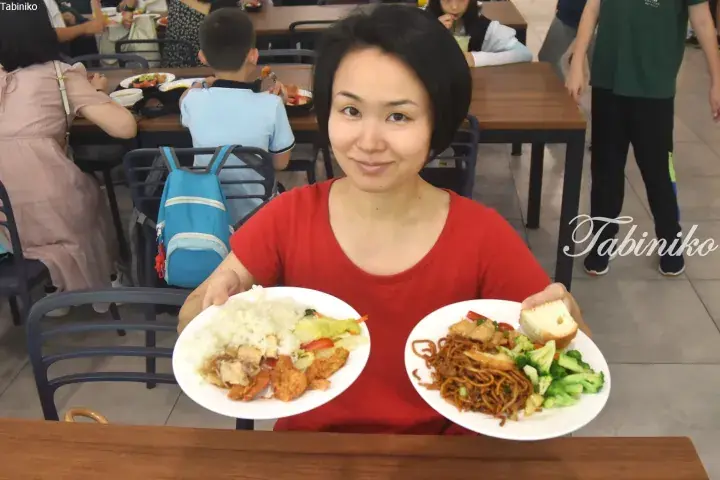 Smiling woman in a red shirt holding two plates of food, including rice and noodles, at a bustling Malaysian food court.