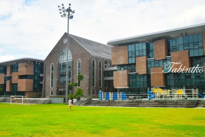 Modern brick buildings with large windows and a church-style structure beside a green lawn with playground equipment at a Malaysian campus.