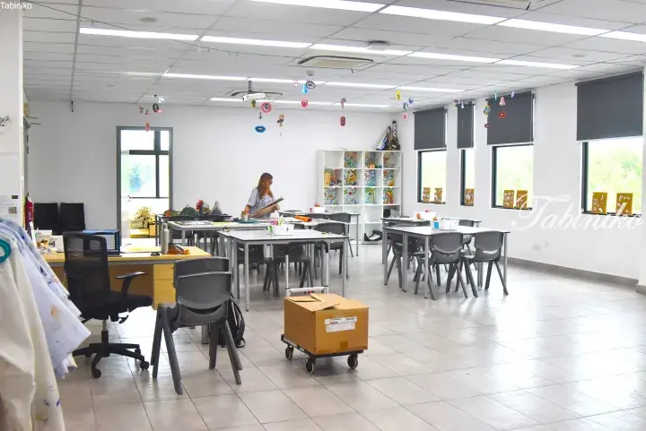 Bright art classroom with tables, chairs, and student artwork displayed on shelves and hanging from the ceiling.