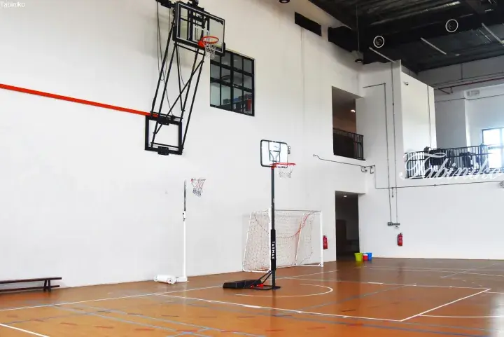 Indoor basketball court with wall-mounted hoop, portable Tarmak hoop, and soccer goal in a spacious gymnasium.