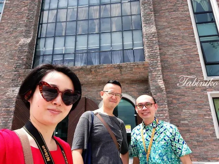 Three tourists smile in front of a brick building with large windows at a heritage site in Malaysia.