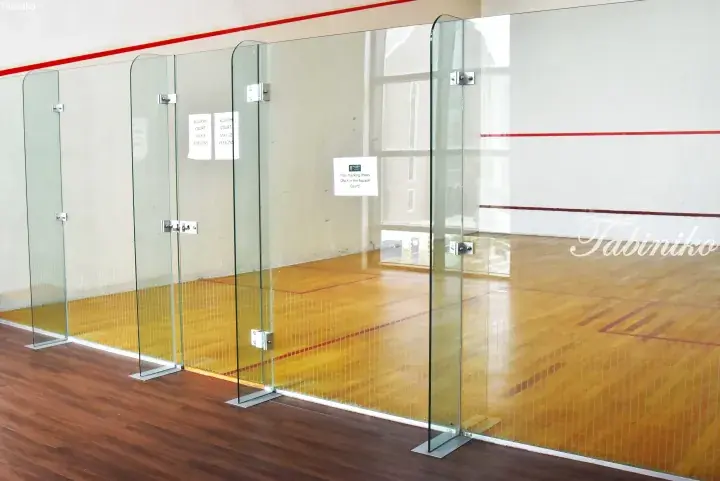 Modern squash court with glass walls, wooden flooring, and red boundary lines, featuring a non-marking shoes sign.