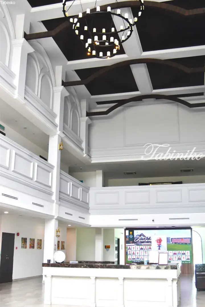 Grand white lobby with high ceilings, dark wood beams, and a modern chandelier above a marble reception desk.