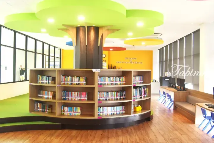 Colorful tree-shaped bookshelf in KHB Primary Library, featuring bright green ceiling accents and organized children&rsquo;s books.