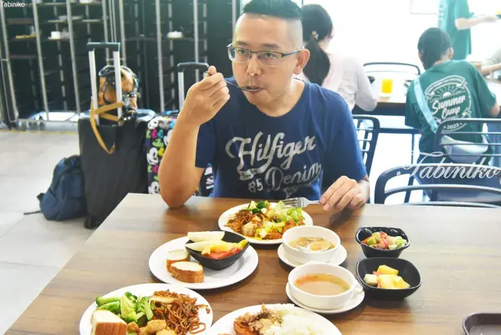 Man eating buffet lunch at airport lounge with luggage nearby, featuring noodles, rice, soup, and fruit.
