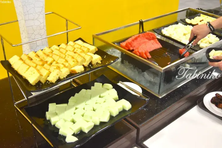 Buffet display featuring sliced sponge cake, green jelly cubes, watermelon, and melon on black trays with ice.