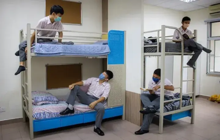 Students in uniform relaxing in a dormitory with bunk beds, reading and resting while wearing face masks.