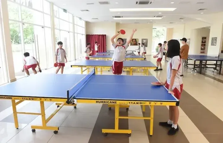 Students celebrating a point during a table tennis match at a bright school sports hall with multiple blue tables.