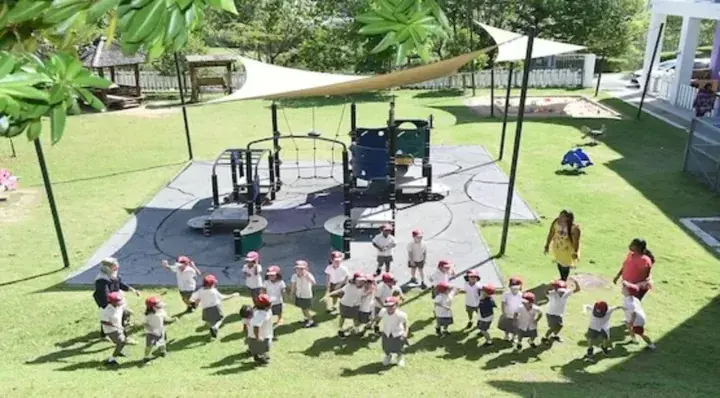 Children in uniform and teachers playing on a playground with shade sails and green grass.