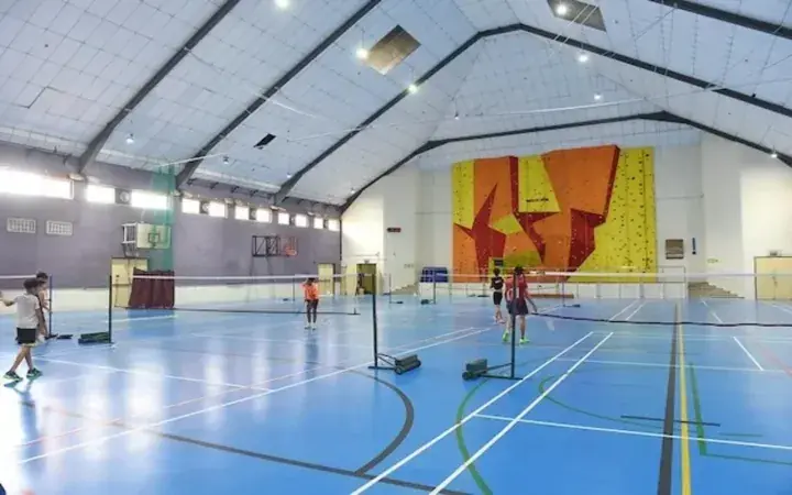 Indoor sports hall with blue flooring, badminton nets, and a colorful climbing wall where children are playing.