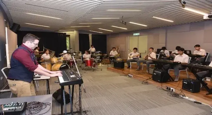 Music students rehearsing with keyboards, drums, and brass instruments in a modern practice room.