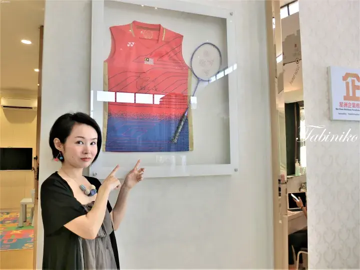 Woman pointing at a framed Lee Chong Wei signed jersey and badminton racket displayed in a Sin Chew Business Excellence office.