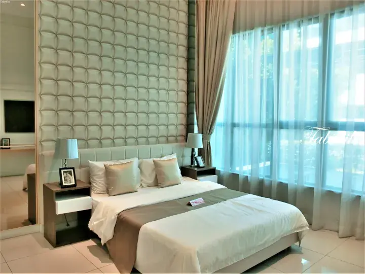 Modern hotel bedroom featuring a large bed with beige tufted headboard and wall paneling, beside a window with sheer curtains.
