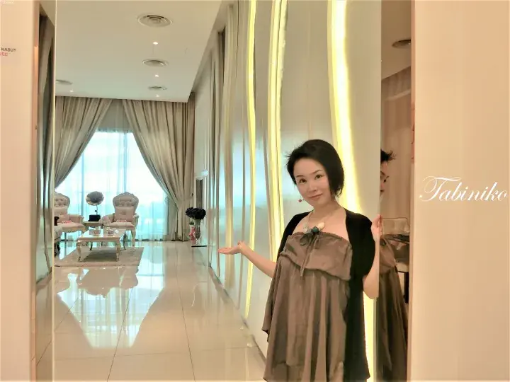 Woman in a brown dress gesturing towards a luxurious hotel suite interior with elegant furniture and large windows.