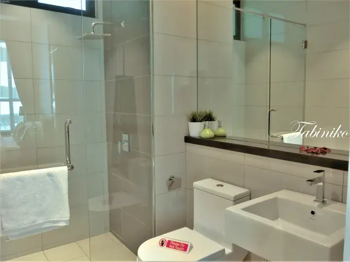 Modern hotel bathroom with glass shower, sink, and toilet featuring a ‘Please Do Not Touch’ sign.