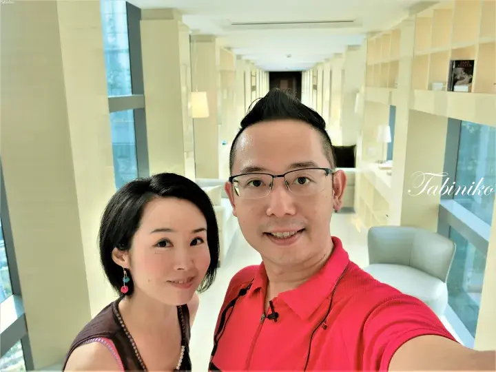 一对夫妇在现代酒店套房的明亮走廊里微笑着自拍，背景是奶油色墙壁和内置置物架。