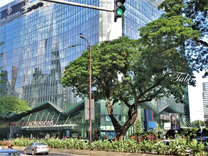 吉隆坡四季酒店与罗宾逊百货大楼的现代玻璃幕墙外观，前景有茂盛的树木。