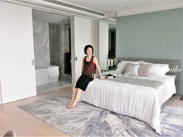 一位女士坐在宽敞的酒店套房内的现代床上，背景可见大理石浴室和淡蓝色墙面。