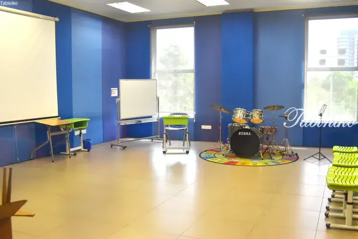Bright music classroom with blue walls, TAMA drum set, whiteboard, and green student desks.