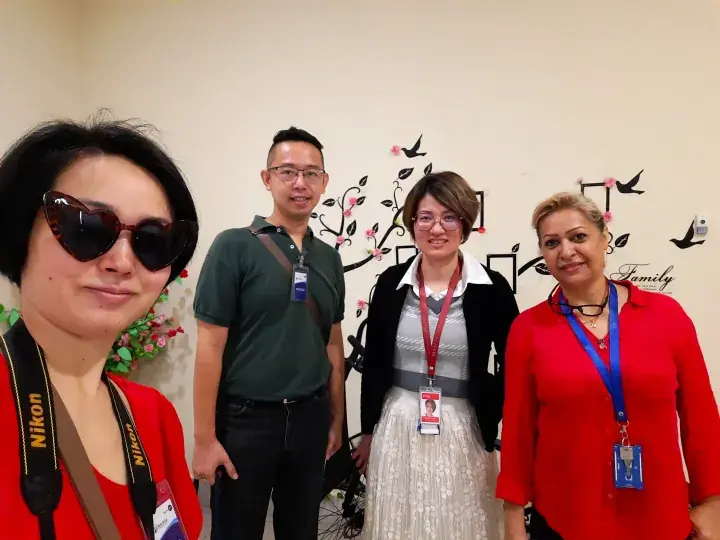 Four people posing together indoors, wearing lanyards and ID badges, with a decorative wall featuring birds and the word ‘Family’ behind them.