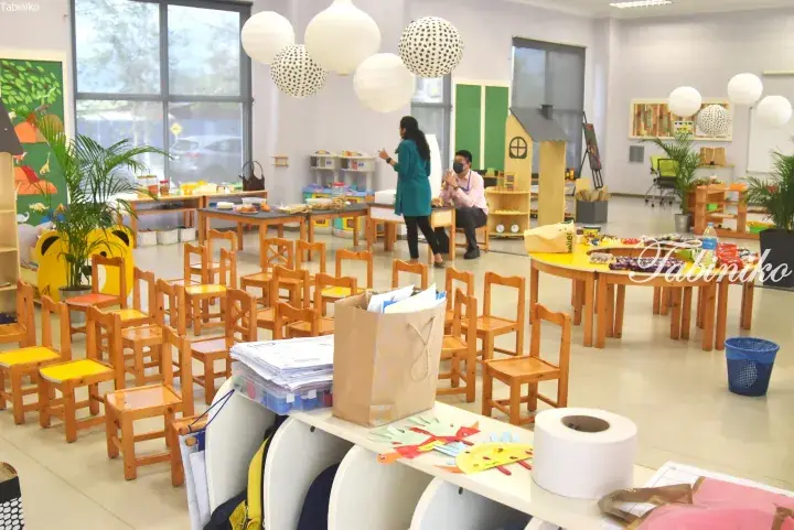 Bright kindergarten classroom with wooden chairs, paper lanterns, and two adults conversing near activity tables.